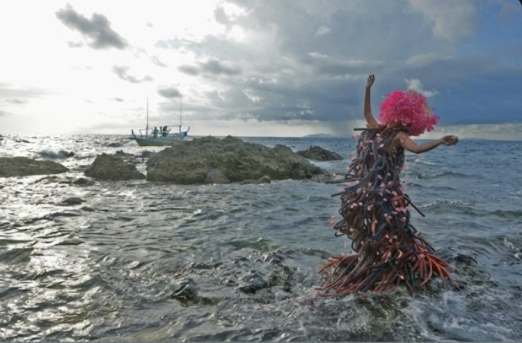 A flyer for an academic book talk titled "Global Asias Book Series II: Filipino Time: Affective Worlds and Contracted Labor (2022) by Allan Punzalan Isaac". The top half of the flyer features a photograph of a performer in the ocean, wearing a large pink wig and a complex costume made of many red, blue, and pink tubes, standing on rocks. A small boat is visible in the distance under a cloudy sky. Below the image, text describes the book's focus on Filipino contract labor and lists the moderator as Andy Urban, with respondents including Christine Bacareza Balance, Lucy M. Burns, Martin Manalansan, Penny Yeung, and Ben Sifuentes.